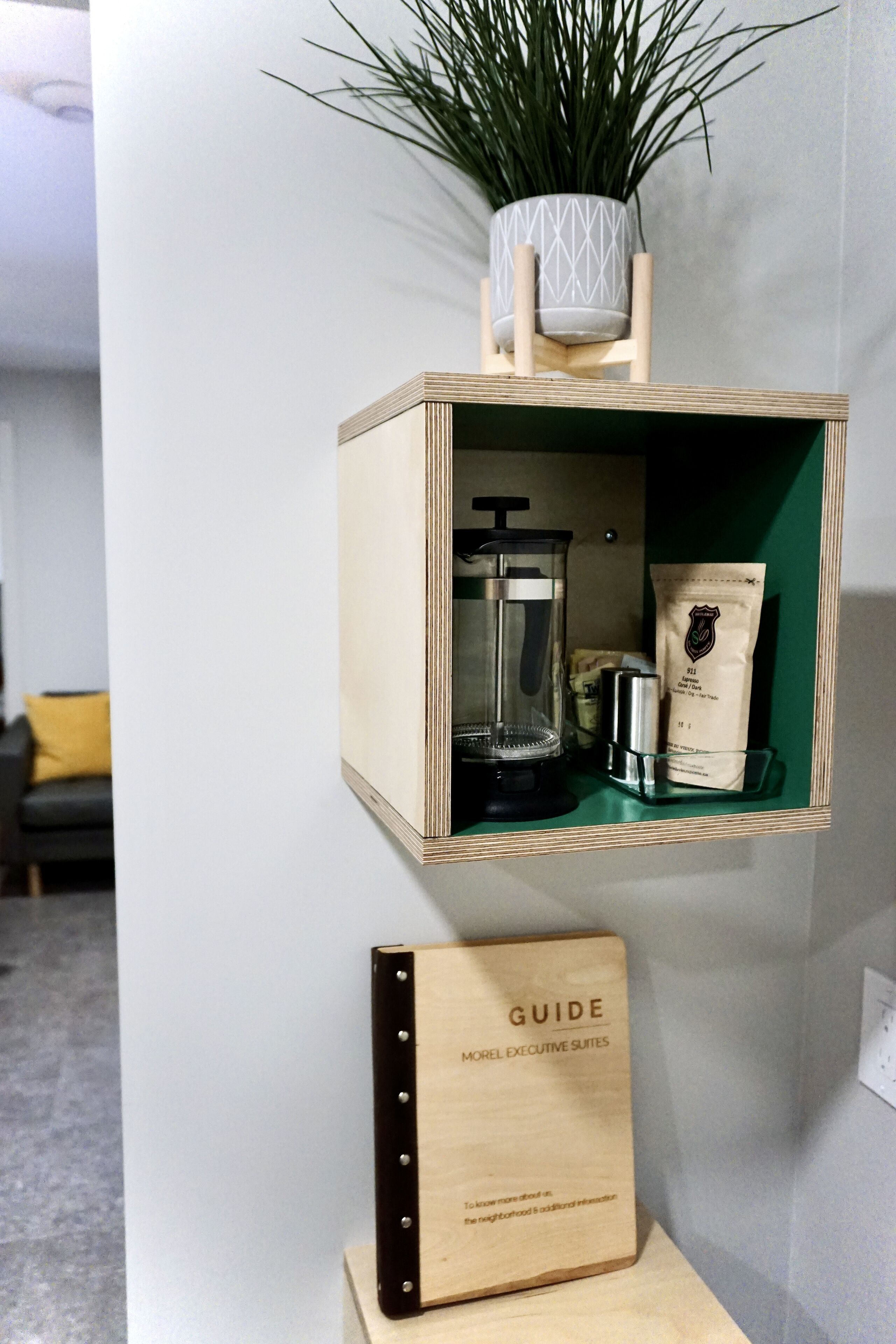 A wooden shelf with a coffee maker, a bag of coffee, and a potted plant. Below the shelf is a wooden plaque with the text "GUIDE Morel Executive Suites".