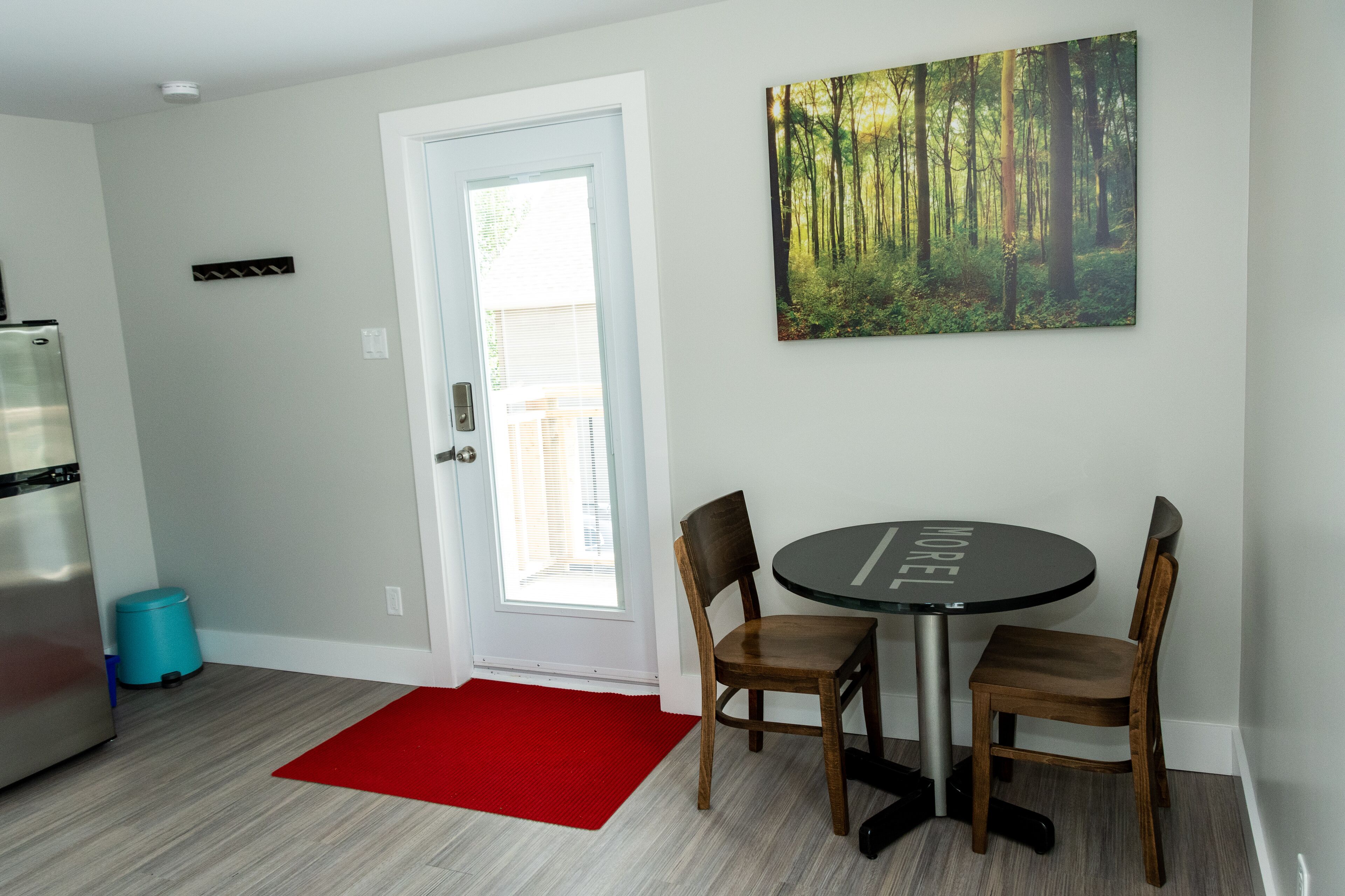 A dining area with a round table and four chairs, a red rug, a refrigerator, and a painting of a forest on the wall.