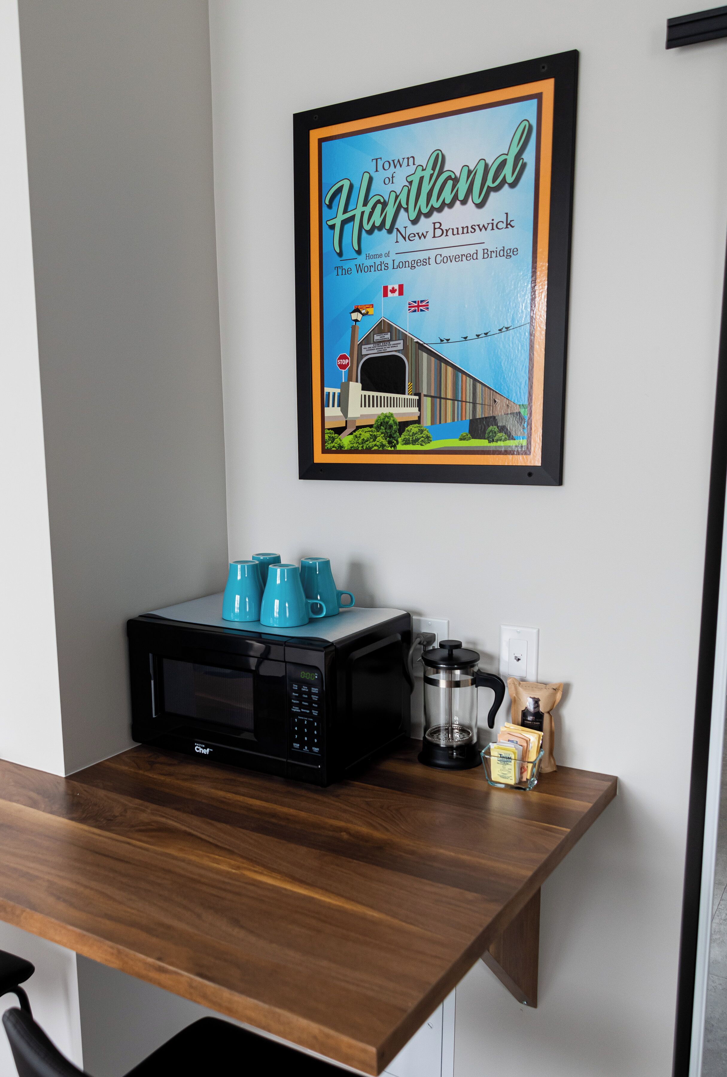 A kitchen area with a microwave, coffee maker, and blue mugs on a wooden countertop. A framed picture of a covered bridge hangs on the wall.