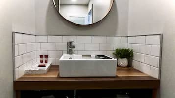 A bathroom with a rectangular sink, a round mirror, white tiles, a small potted plant, and two towels on a shelf.