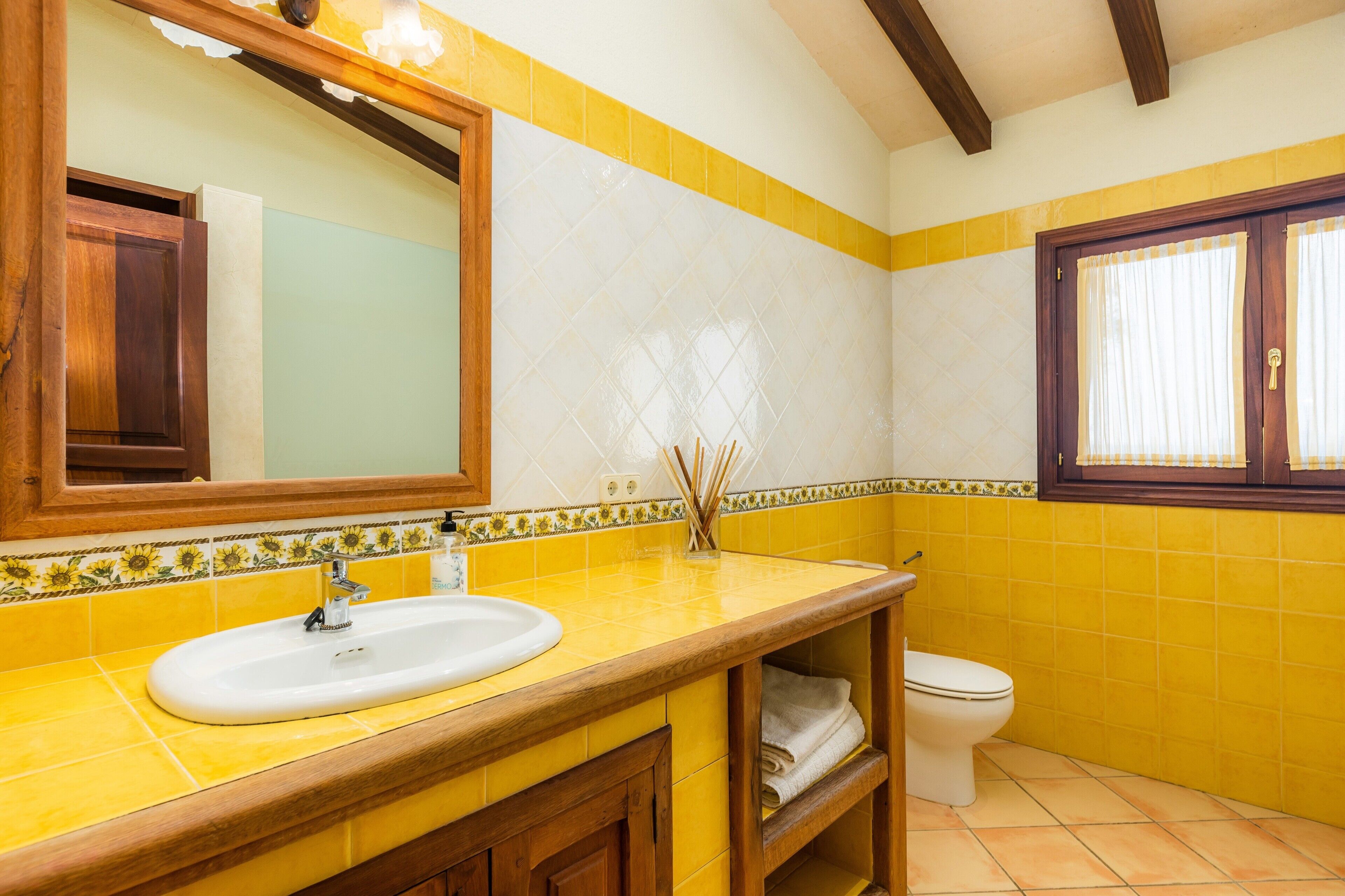 Een badkamer met een wand betegeld in geel en wit, een houten wastafelmeubel met een wastafel, een spiegel en een toilet.