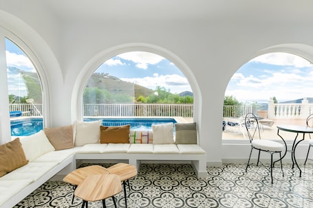 A room with a white sofa, a small wooden table, and a view of a pool and mountains.. Villa "Casa Silencio" with Sea View, Private Pool & Wi-Fi