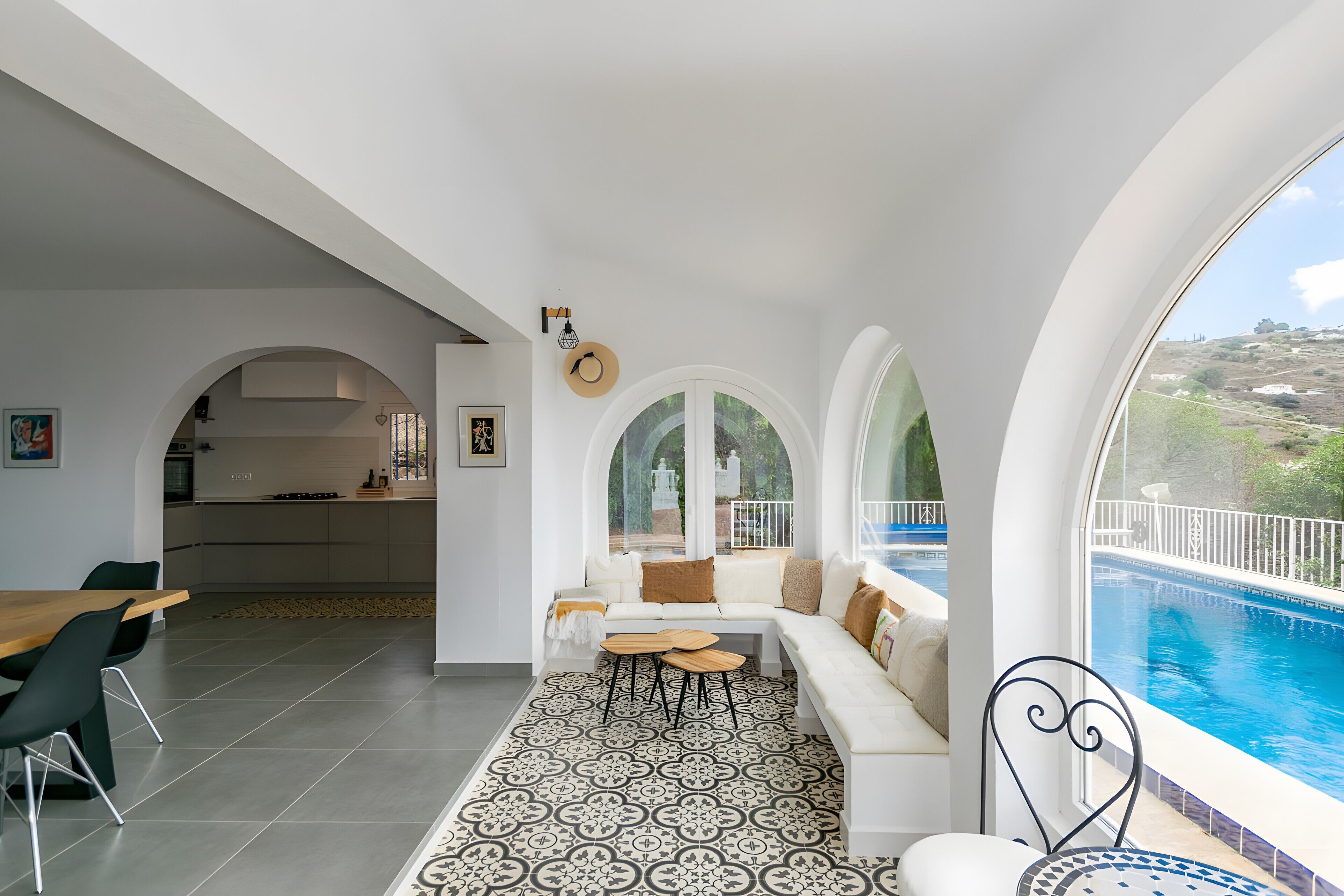 A modern interior with a dining area, a sofa with a view of a swimming pool, and a kitchen visible through an archway.