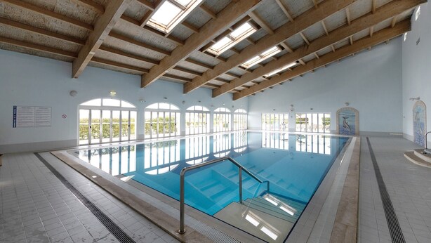 Una piscina interior con suelo de baldosas, vigas de madera y grandes ventanales.