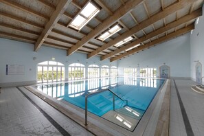 An indoor swimming pool with a tiled floor, wooden beams, and large windows.