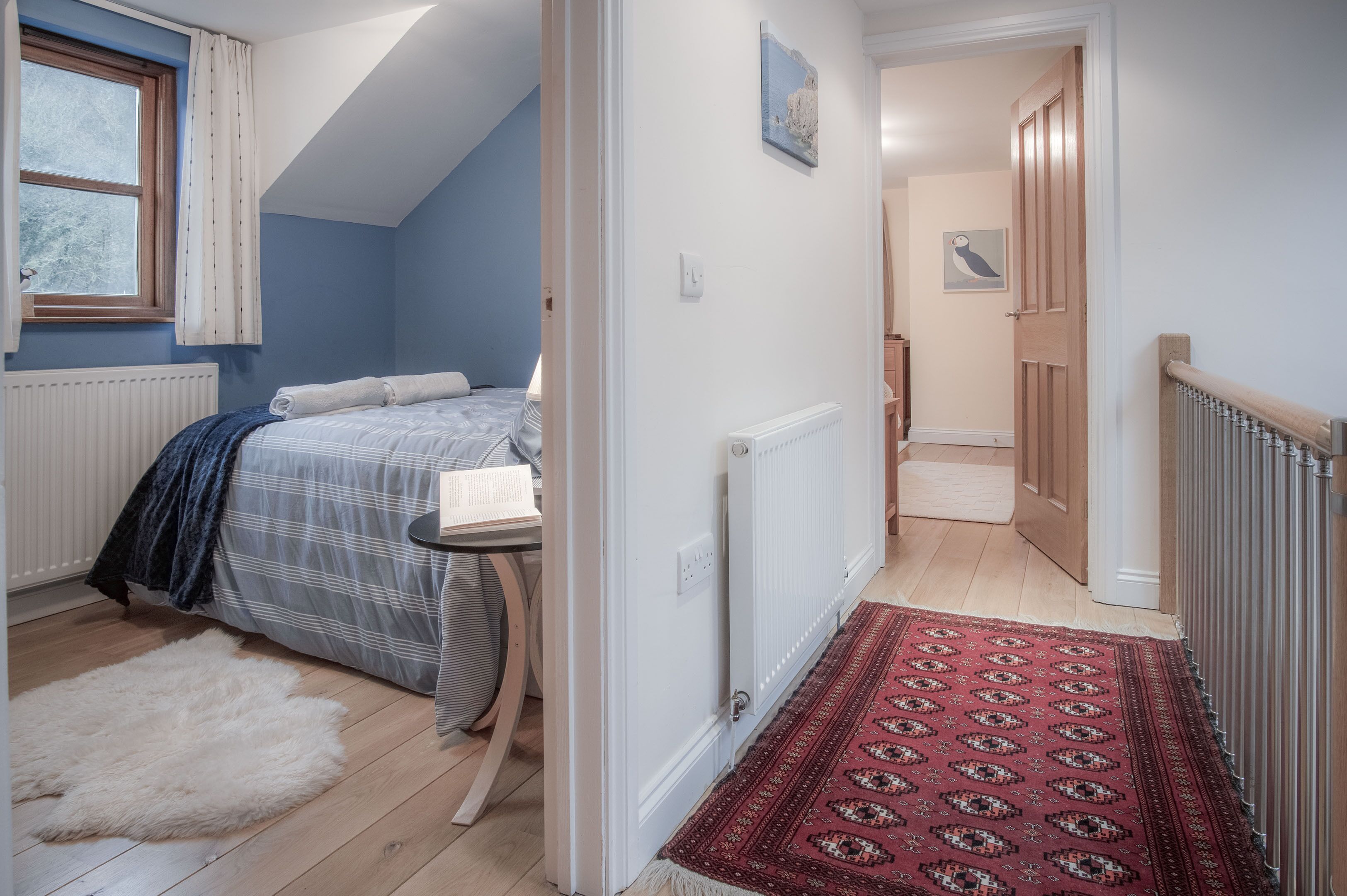 A bedroom with a bed, a small table, a radiator, a window with curtains, and a hallway with a door.