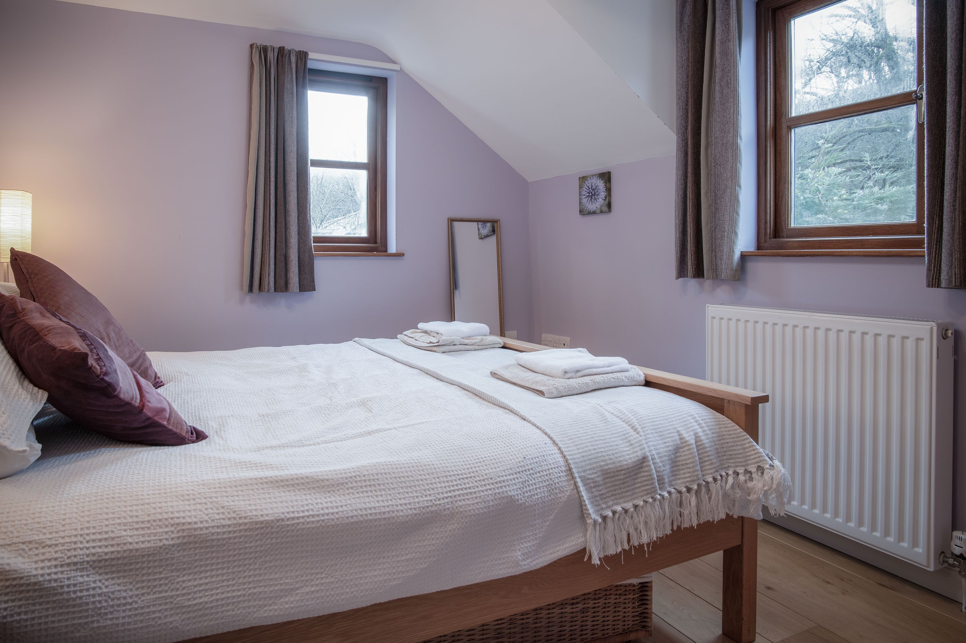 A neatly made bed with a white blanket and pillows, a bedside table with a lamp, and a window with curtains.