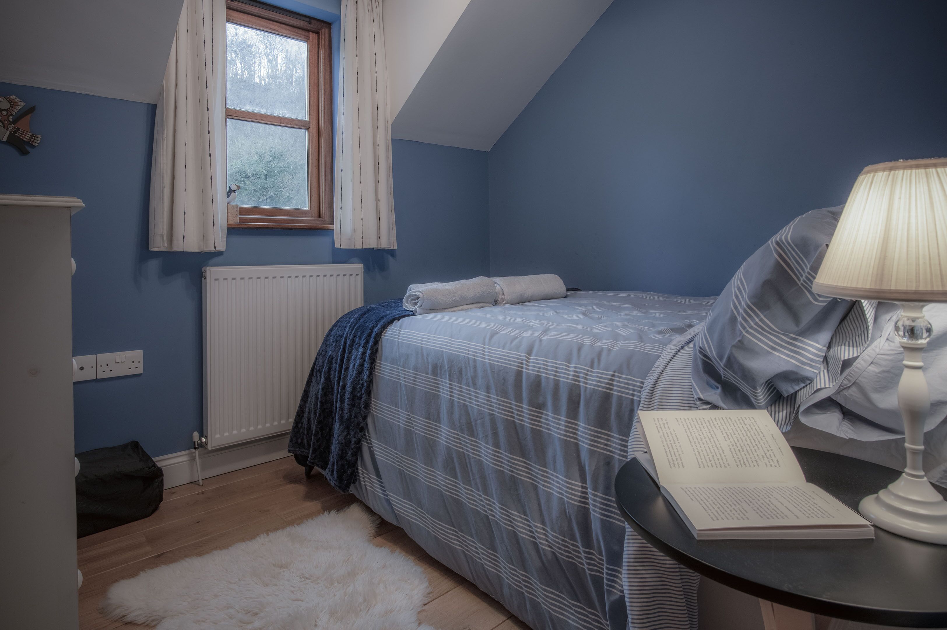 A bedroom with a bed, a bedside table with a lamp, a window with curtains, a radiator, and a white furry rug.