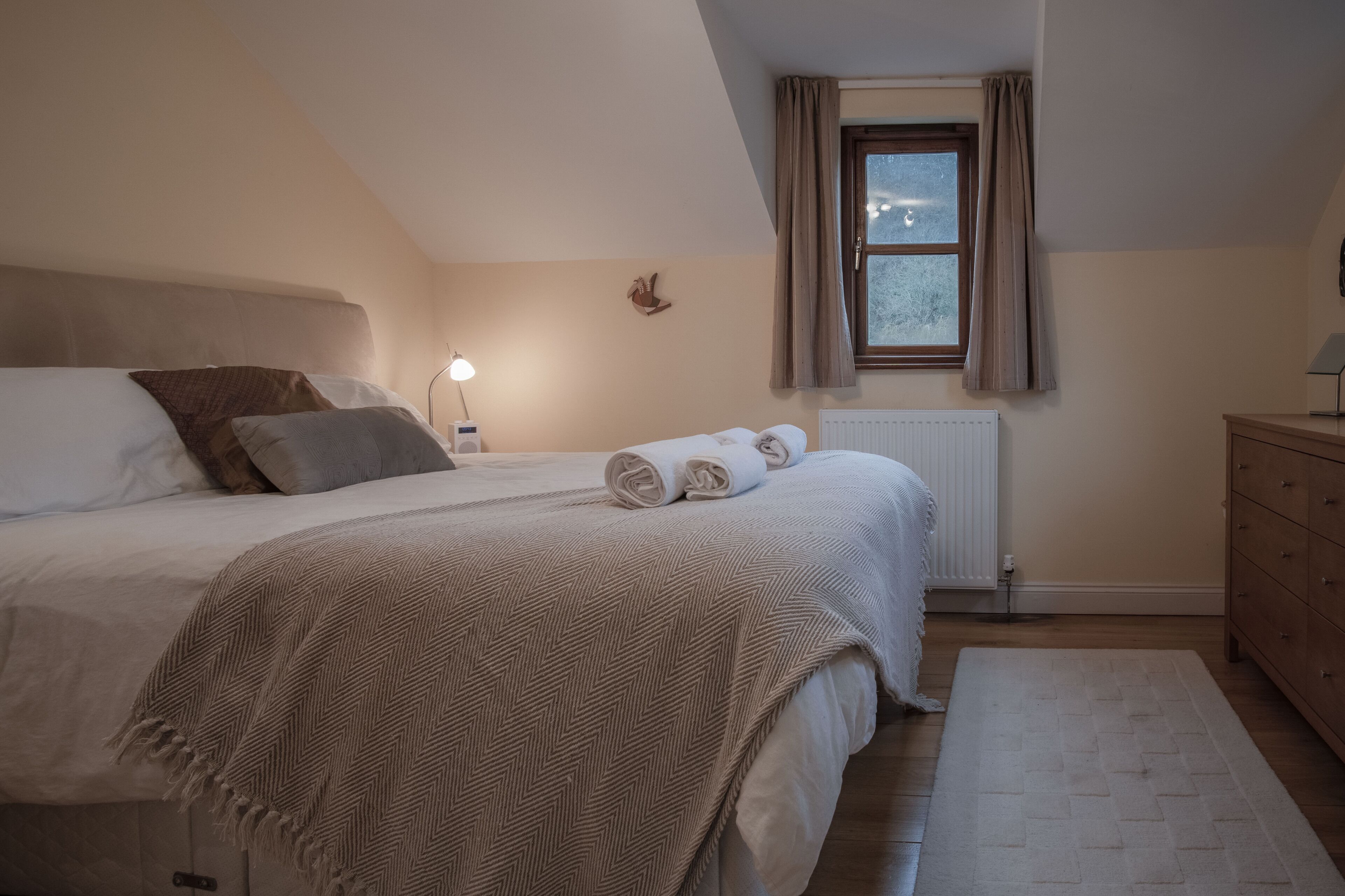 A bedroom with a bed, bedside table, lamp, window, radiator, and wooden dresser.