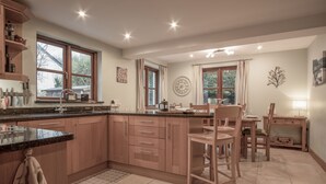 A spacious kitchen with wooden cabinets, a granite countertop, and a dining area with a table and chairs. - Llais Afon - 3 Bedroom Holiday Home - Fishguard (Fishguard)
