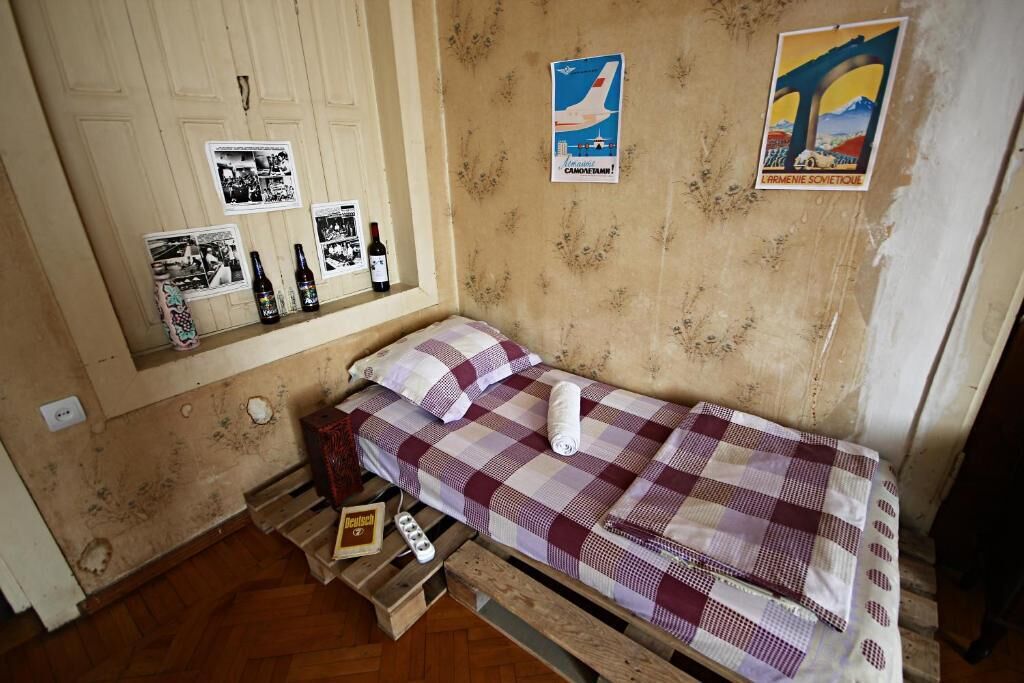 A small, compact room with a single bed, a wooden floor, and a wall with posters and pictures.