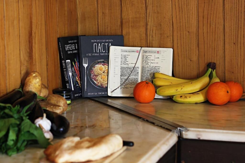 A kitchen counter with a cookbook, a recipe book, and fresh produce including bananas and oranges.