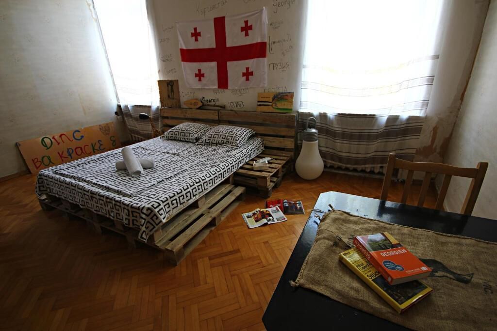 Una habitación con una cama sobre un palet de madera, una bandera en la pared y una mesa con libros.