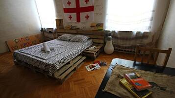 Una habitación con una cama sobre un palet de madera, una bandera en la pared y una mesa con libros.