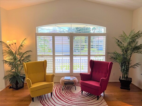 A room with a large bay window, two chairs, a small round table, and two potted plants.