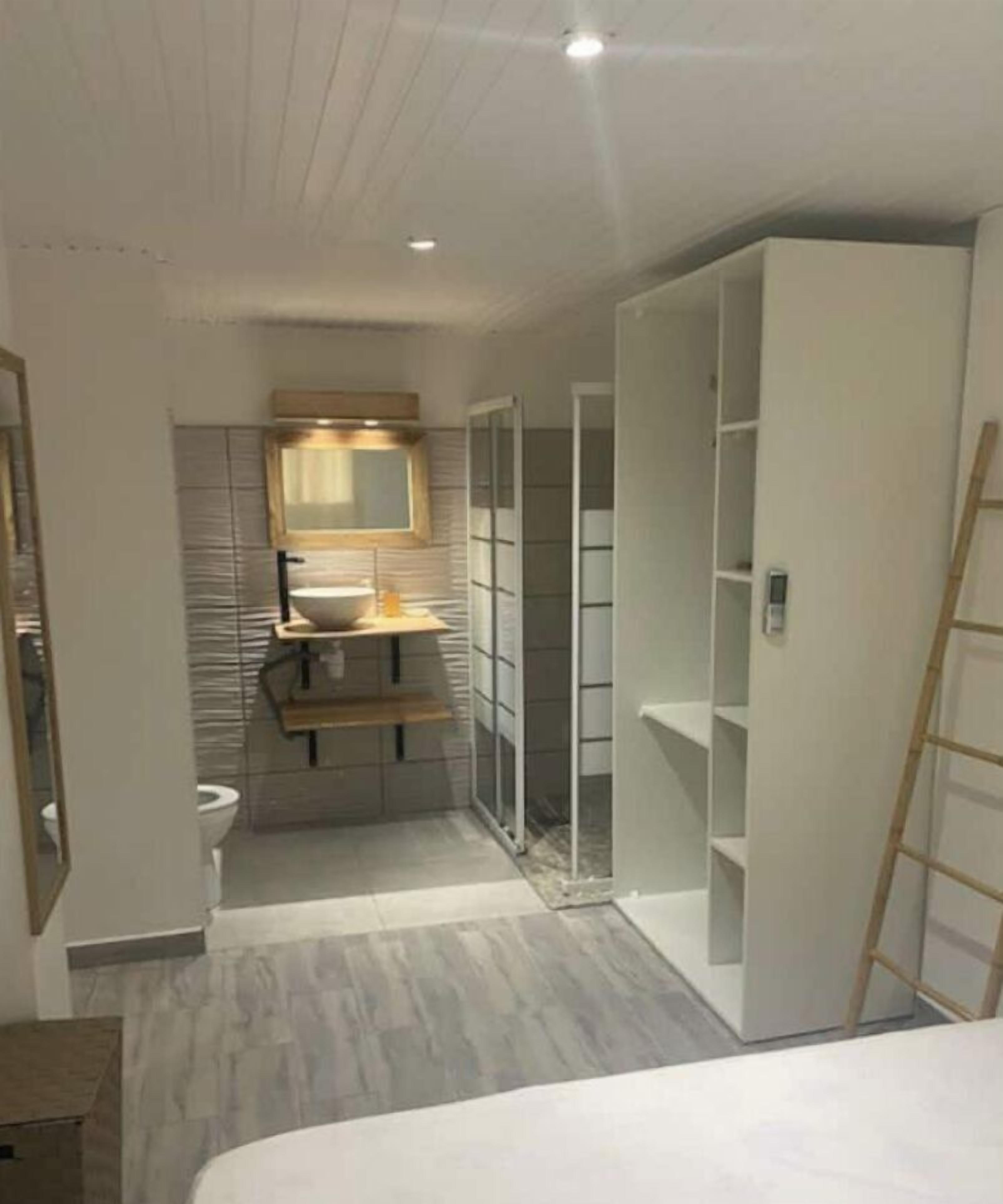 A narrow, modern bathroom with a toilet, sink, and mirror. There are built-in shelves and a ladder leaning against the wall.