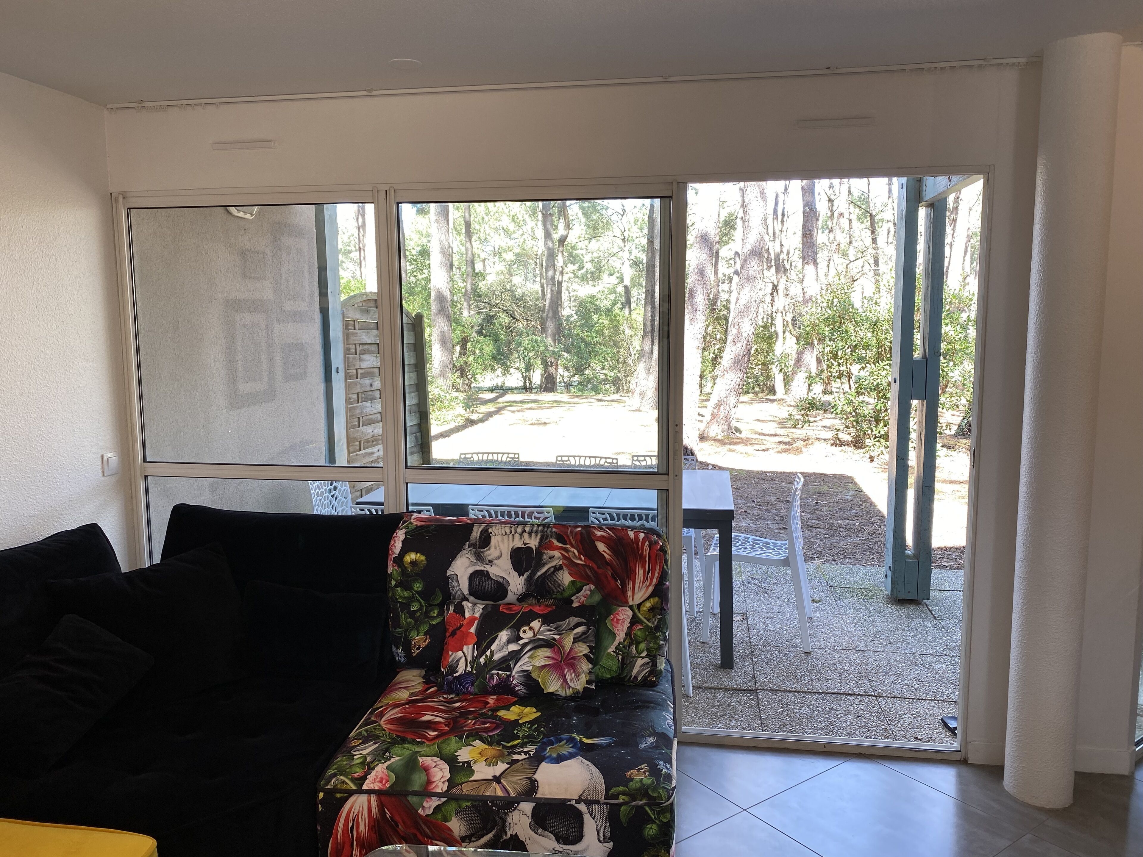 A room with a black couch, a floral-patterned chair, and a view of a forest through large windows.