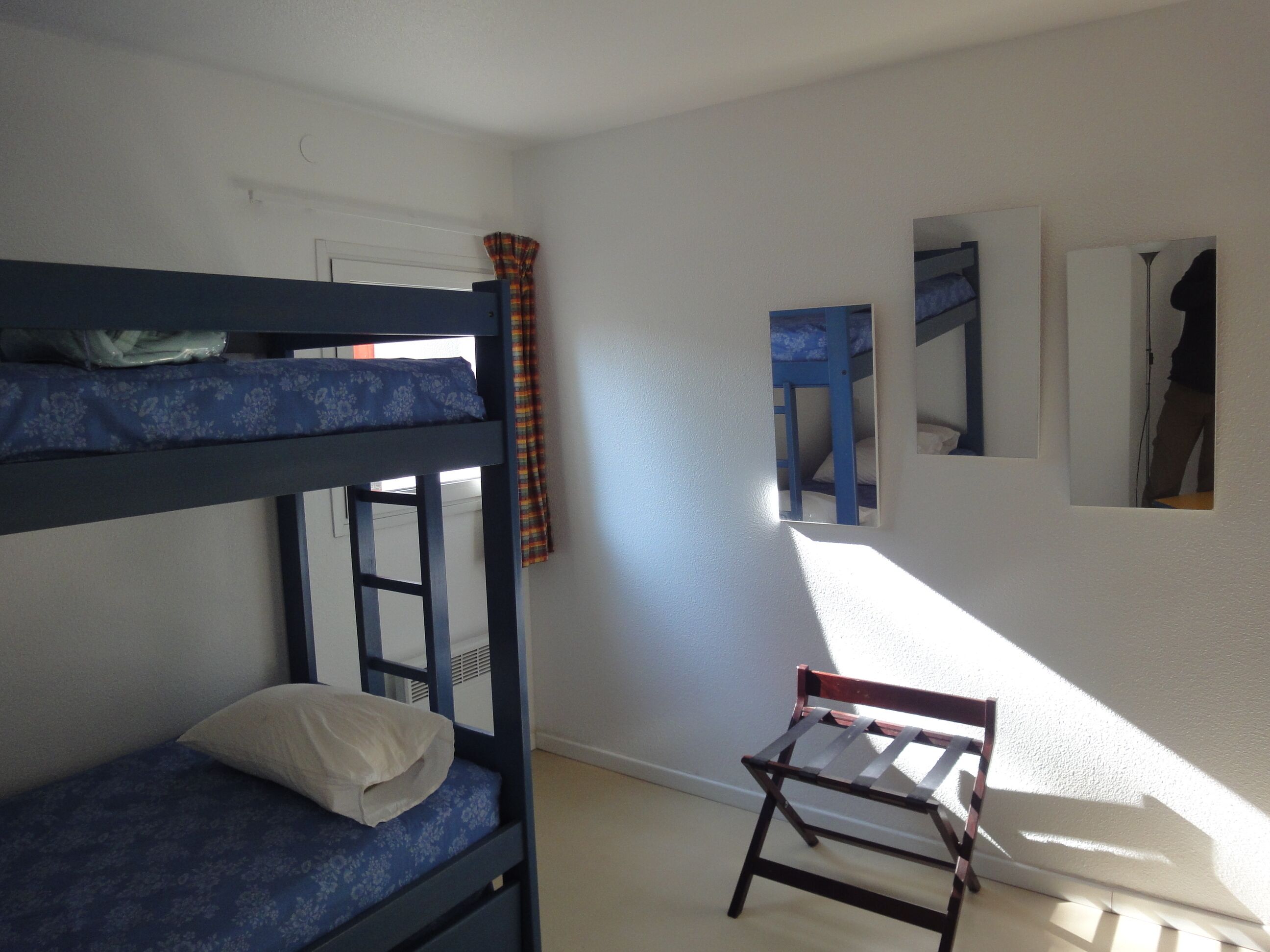 A dormitory room with bunk beds, a small wooden chair, and a window with curtains.