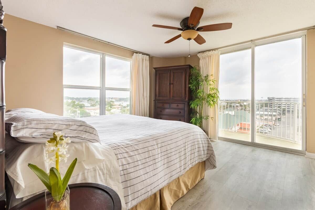 A bedroom with a large bed, a ceiling fan, a view of the city through large windows, and a plant.