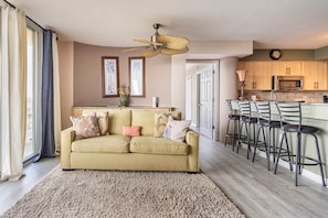 Una sala de estar con un sofá, una mesa de centro y una alfombra. Hay una zona de cocina con un microondas y un reloj.