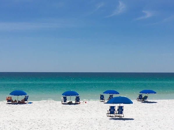 Una playa de arena blanca y agua turquesa cristalina. Varias sombrillas azules ofrecen sombra.