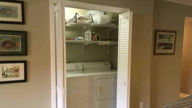 Une buanderie équipée d’un lave-linge et d’un sèche-linge, d’une étagère où sont rangés divers objets, et d’une porte donnant sur une autre pièce.