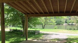 Un espace pique-nique abrité, doté d’un sol en béton, entouré de pelouse et d’arbres, et offrant une vue sur un lac.