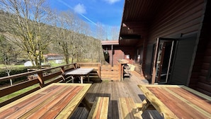 Un pontile in legno con tavoli da picnic e panchine, una zona coperta con tavolo e sedie, e vista su una zona boschiva.