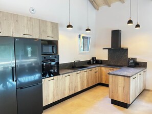 Una cocina moderna con armarios de madera, un refrigerador de acero inoxidable, horno empotrado, microondas, cafetera e iluminación colgante.