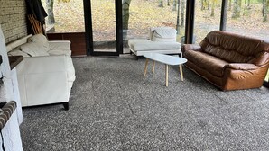 A cozy room with a white sofa, a brown leather couch, and a small table. Large windows offer a view of a wooded area. - Ferme des Jonquilles à Gérardmer (15p) (Gérardmer)