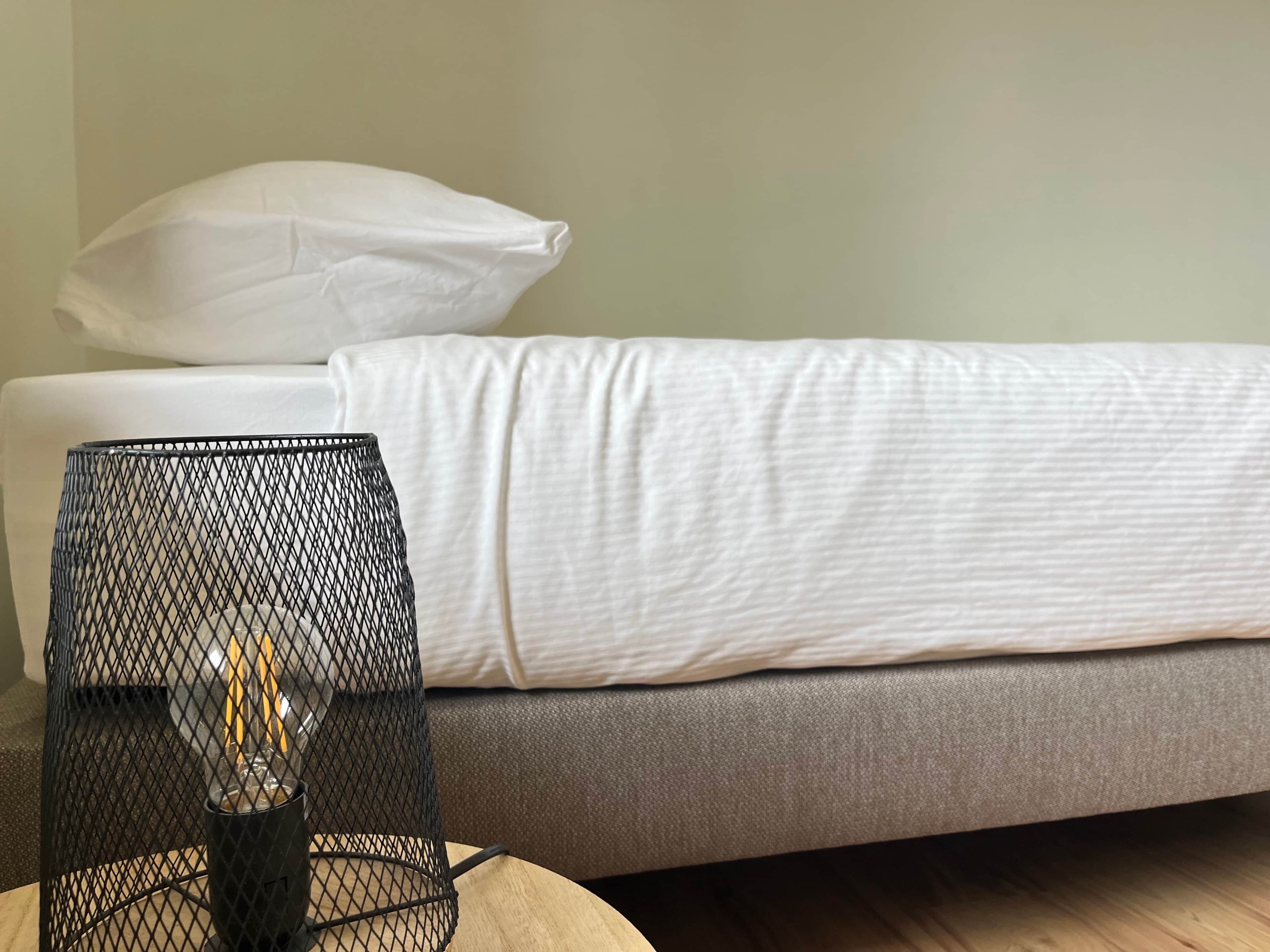 Una cama con ropa de cama blanca, un sofá gris y una lámpara de malla negra sobre una mesa de madera.