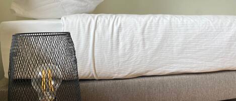 Una cama con ropa de cama blanca, un sofá gris y una lámpara de malla negra sobre una mesa de madera.