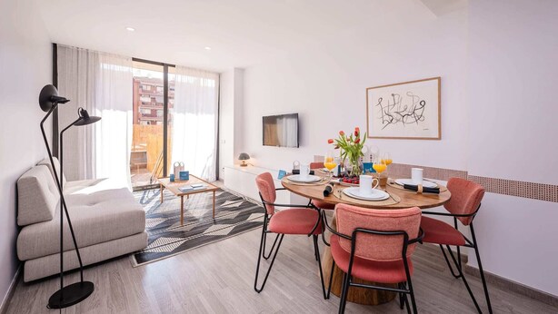 A modern living room with a sofa, a dining table with chairs, a TV mounted on the wall, and a large window with curtains. - Sweett | Fira 104 - Two Bedroom Apartment, Sleeps 4 (Barcelona)