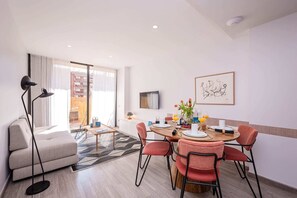 A modern living room with a sofa, a dining table with chairs, a TV mounted on the wall, and a large window with curtains. - Sweett | Fira 104 - Two Bedroom Apartment, Sleeps 4 (Barcelona)
