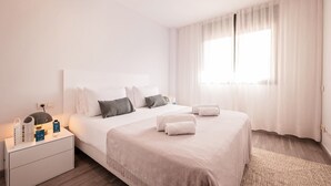 Une chambre d’hôtel moderne avec un grand lit, une table de chevet avec une lampe et une fenêtre ornée de rideaux légers.