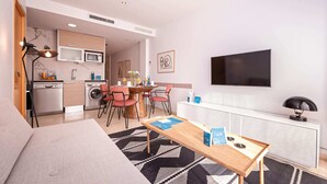 Sala de estar moderna com sofá, mesa de centro de madeira e área de refeições com mesa e cadeiras. Há uma cozinha com micro-ondas e máquina de lavar. Uma TV está fixada na parede e há uma janela com persianas.