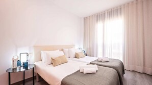 Een hotelkamer met twee bedden, een klein tafeltje met een lamp en een raam met gordijnen.