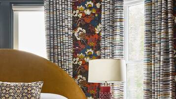 A neatly made bed with a patterned pillow, a small bedside table with a lamp and a plant, and a window with curtains.