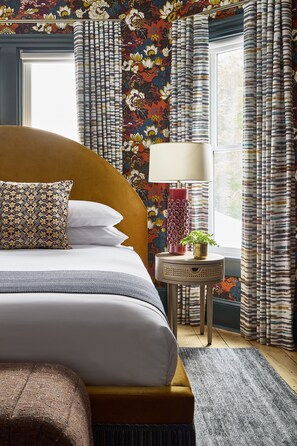 A neatly made bed with a patterned pillow, a small bedside table with a lamp and a plant, and a window with curtains.