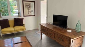 Una habitación con un mueble bajo de madera para el televisor, un sofá amarillo con cojines oscuros, una mesa de madera y un cuadro en la pared.