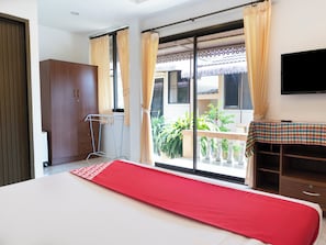 Relax in this inviting room with a large bed, a wardrobe, and a table with a checkered tablecloth, plus a TV and sliding glass door to a balcony with potted plants.