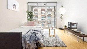 Une chambre moderne avec un lit, une télévision, une petite table et une grande fenêtre donnant sur des immeubles.