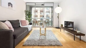 Un salon moderne avec un canapé noir, une petite table en bois, un téléviseur posé sur un meuble et une grande fenêtre donnant sur des immeubles.