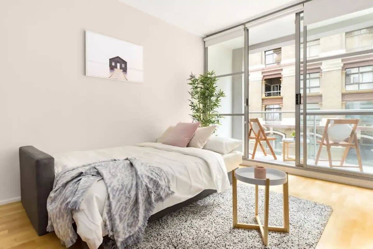 A modern bedroom with a bed, a small round table, a large window with a view of buildings, and a potted plant.