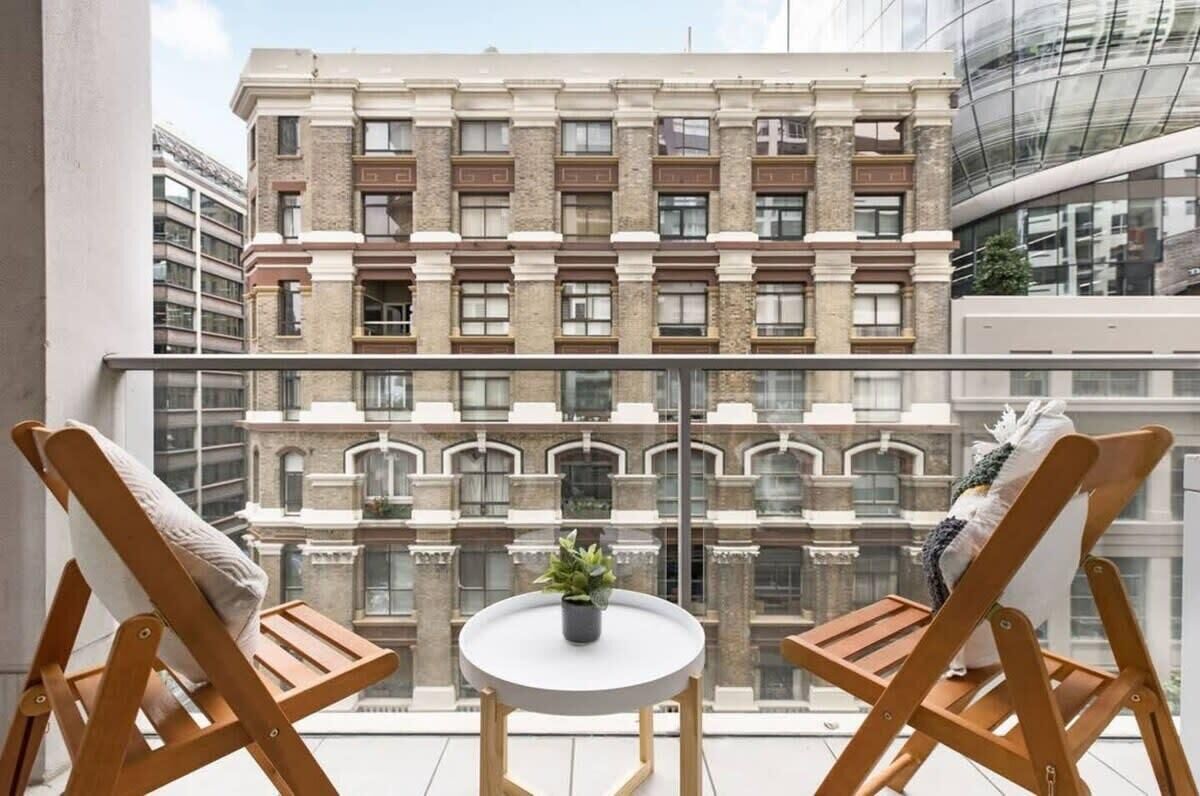 A balcony with two wooden chairs and a small round table with a plant, overlooking a historic building with multiple windows and balconies.