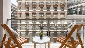 Un balcon avec deux chaises en bois et une petite table ronde agrémentée d’une plante, donnant sur un bâtiment historique doté de nombreuses fenêtres et de balcons.