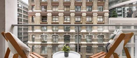 Un balcón con dos sillas de madera y una pequeña mesa redonda con una planta, con vista a un edificio histórico con múltiples ventanas y balcones.