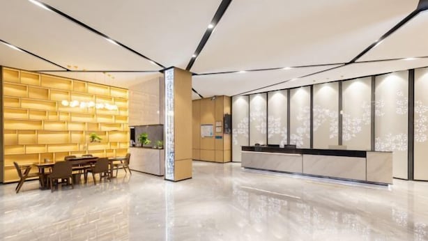 A modern hotel lobby with a wooden reception desk, a seating area with tables and chairs, and a large decorative wall with vertical panels. - Langfeng Capital Hotel Zhongshan (Former Sun Yat-sen · Nanlang Station Branch) (Zhongshan)