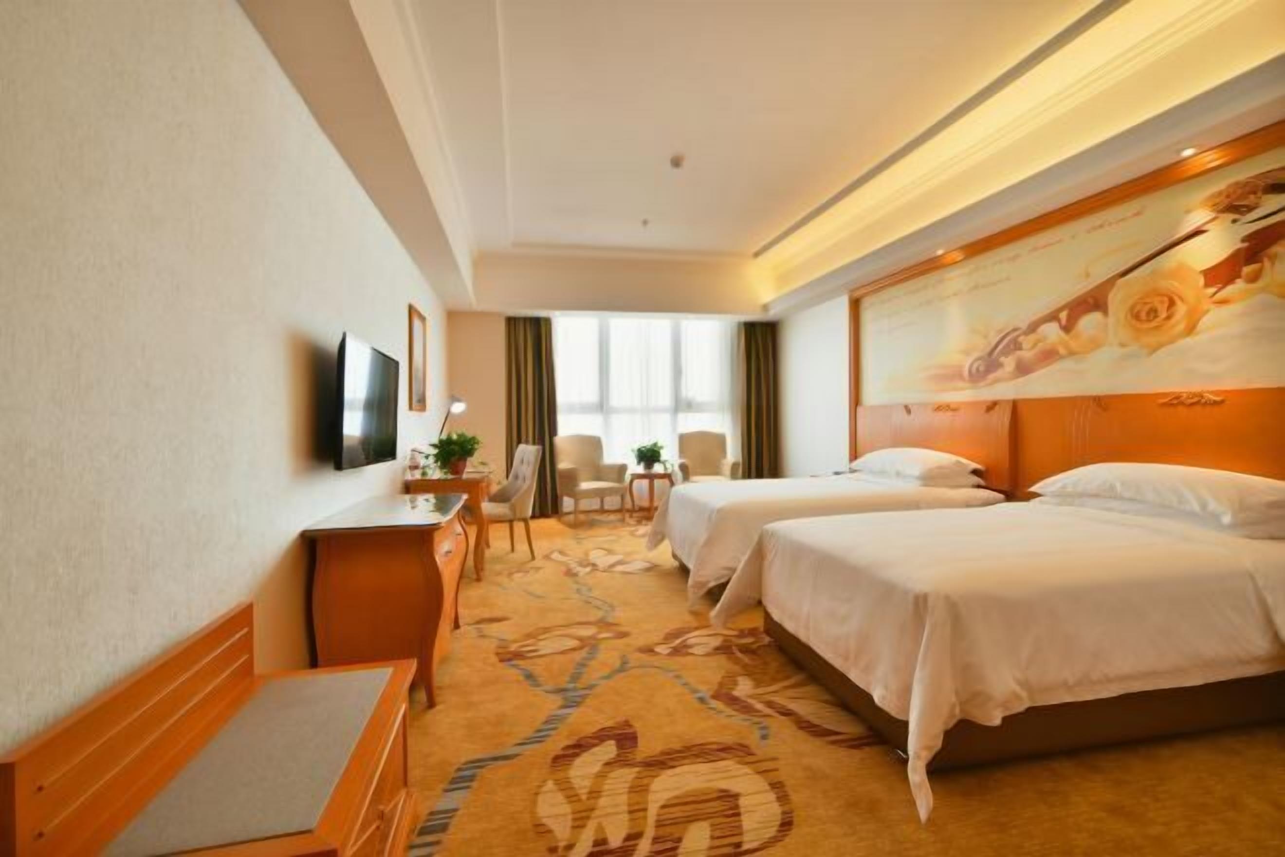 A hotel room with two beds, a desk with a chair, a TV, and a window with curtains.