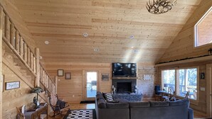 A wooden cabin interior with a large gray sofa, a fireplace, a TV, and a staircase. - Beautiful New Cabin on the Broad River, 3 miles from Lake Lure, NC (Lake Lure)